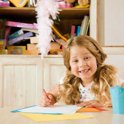 A young girl with curly blonde hair is smiling while holding a red pencil, engaged in coloring with kids, a fun fine motor activity at a table with colorful papers in a creative playroom.